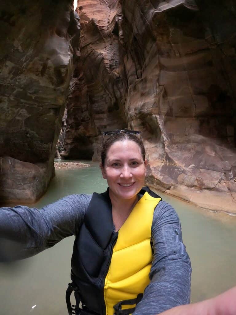 Selfie of me in a yellow life jacket, standing in water up to my waist, with the beautiful canyon of Wadi Mujib in the background.