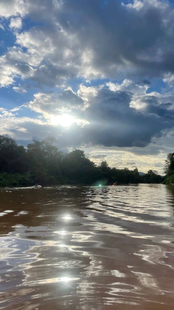 View of the Ping River in Chiang Mai from a kayak on the water. Clouds and sun in the distance.