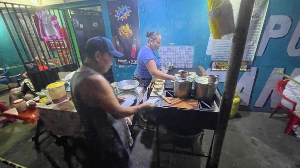 Two cooks prepare traditional Salvadoran dishes at an outdoor eatery with a blue-painted wall. The woman in the foreground is cooking tortillas on a flat griddle, while the man beside her handles other ingredients. Large pots and utensils are visible on the cooking station, with a menu and advertisement poster on the wall behind them.