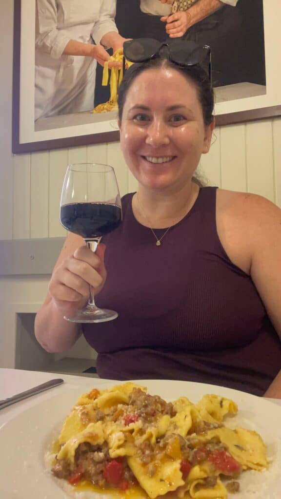 Smiling solo female traveler (me) holding a glass of red wine while seated in front of a plate of freshly made pasta with meat and tomato sauce, inside a restaurant in Parma, Italy.
