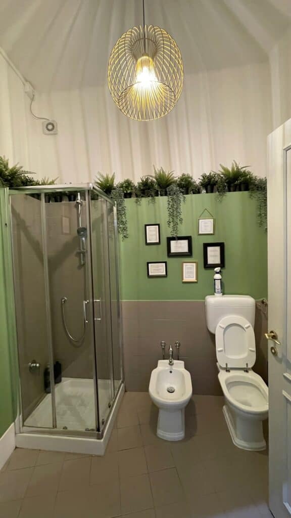 Modern bathroom at The Host in Parma, Italy, with a glass-enclosed shower, toilet, bidet, and green plants decorating the wall above framed certificates.