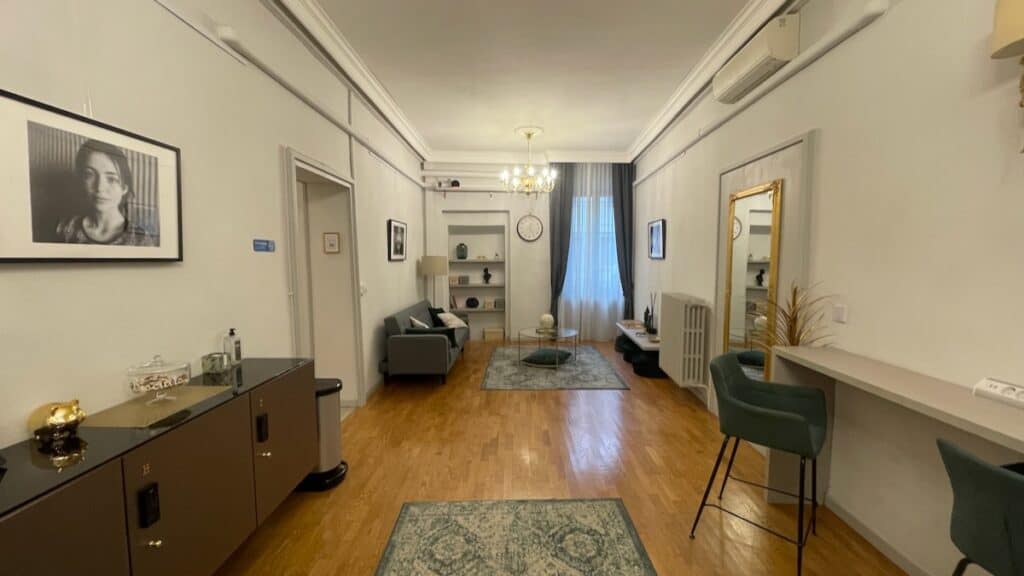 Spacious common area at The Host hostel in Parma, Italy, featuring a cozy seating arrangement with a sofa, wall shelves, and a decorative chandelier hanging from the ceiling.