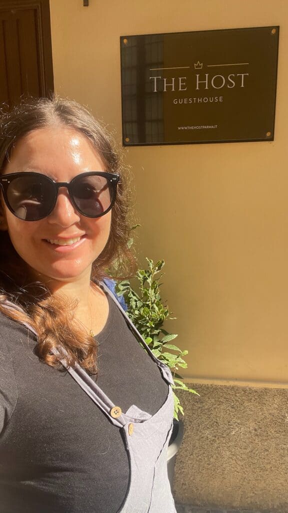 A solo female traveler (me) smiling and wearing sunglasses outside The Host hostel in Parma, Italy, standing in front of a sign that reads 'The Host Guesthouse'